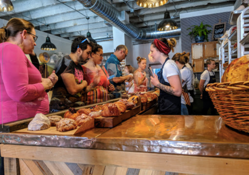 A busy bakery scene with customers in line and a staff member serving pastries at the counter.