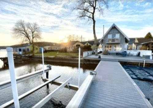 A serene lakeside view at sunset, featuring a wooden dock and a cozy house in the background.