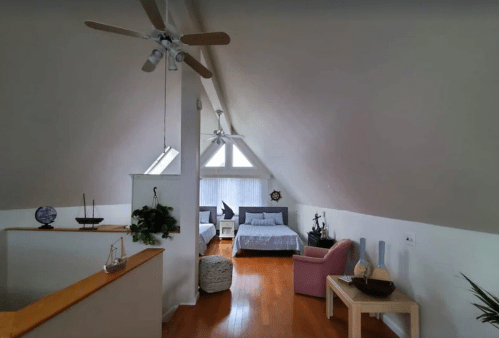 A cozy attic room with two beds, a pink chair, and large windows letting in natural light.