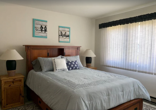 A cozy bedroom featuring a wooden bed, decorative pillows, two lamps, and artwork on the walls. Natural light from a window.