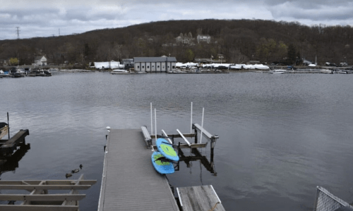 A calm lake scene with a dock, two kayaks, and a hillside in the background under a cloudy sky.