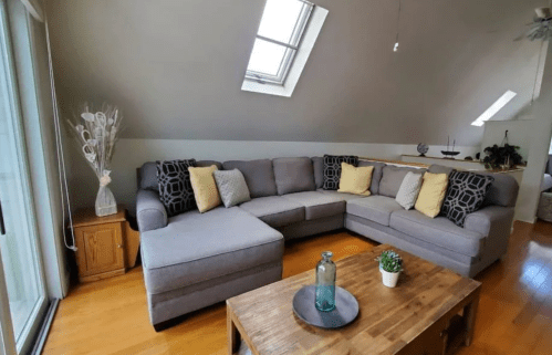 Cozy living room with a gray sectional sofa, yellow pillows, wooden coffee table, and natural light from skylights.