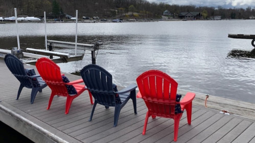 Three colorful Adirondack chairs, two red and one navy blue, sit on a dock overlooking a calm lake.