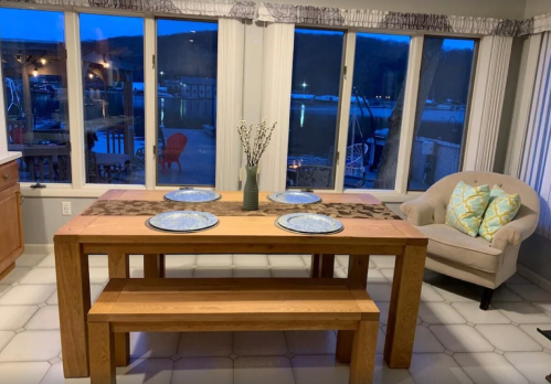 A wooden dining table with plates set, near large windows overlooking a marina at dusk. A cozy chair is nearby.