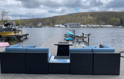A waterfront view featuring a patio set with blue cushions on a dock, overlooking a calm lake and boats.