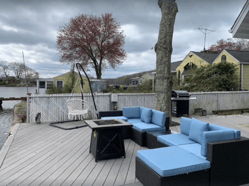 A cozy outdoor seating area with blue cushions, a fire pit, and a view of a lake under a cloudy sky.