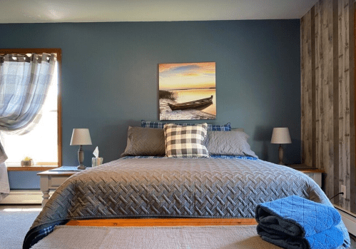 Cozy bedroom with a blue accent wall, a large bed, plaid pillows, and a scenic painting above the headboard.