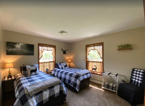 Cozy bedroom with two plaid beds, warm lighting, and rustic decor, featuring windows with nature views.