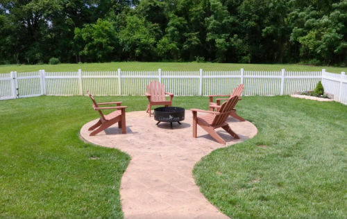 A cozy outdoor seating area with four wooden chairs around a fire pit, surrounded by a green lawn and white fence.