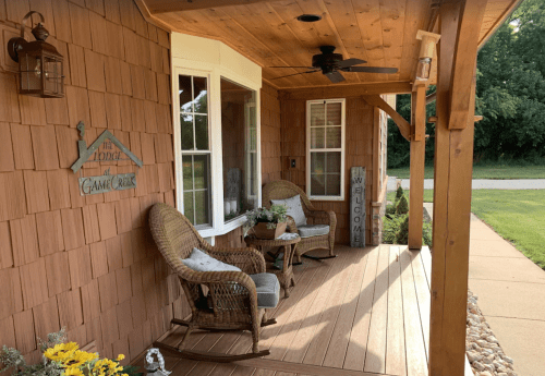 A cozy porch with two wicker chairs, a small table, and welcoming decor, surrounded by greenery.