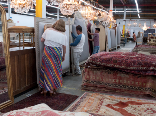 People rearranging large rugs and mirrors in a spacious store filled with chandeliers and furniture.