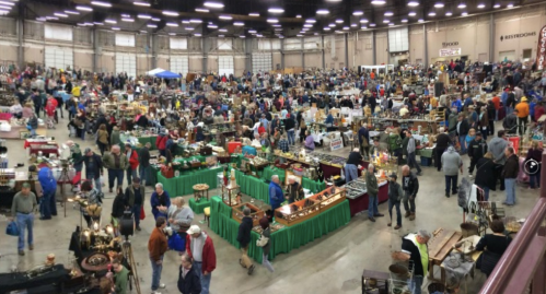 A bustling indoor market filled with vendors and crowds browsing various antiques and collectibles.