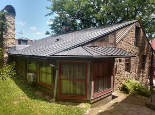 A rustic stone house with a metal roof, surrounded by greenery and a clear blue sky.
