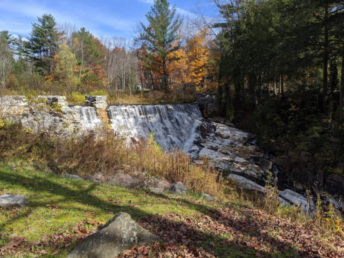 A serene waterfall cascades over rocky ledges, surrounded by autumn foliage and trees under a clear blue sky.