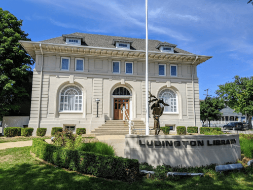 Historic Ludington Library building with a statue in front, surrounded by greenery and a clear blue sky.