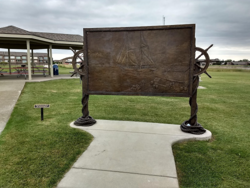 A large bronze relief sculpture depicting a sailing ship, set in a park with a gazebo in the background.