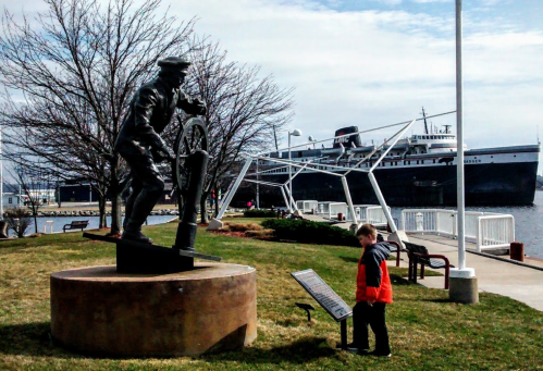 A child observes a statue of a sailor at a waterfront park, with a ship docked in the background.