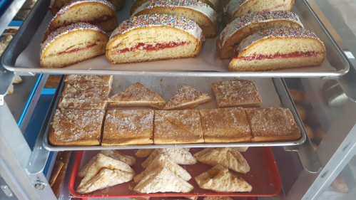 A display of baked goods: colorful frosted cakes with sprinkles, triangular pastries, and square cookies on trays.