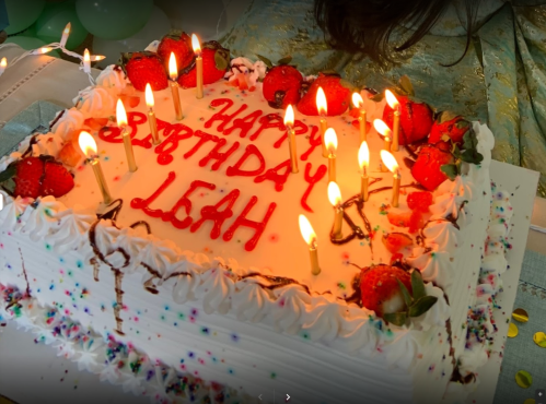 A birthday cake with "Happy Birthday Leah" written in red icing, topped with strawberries and lit candles.