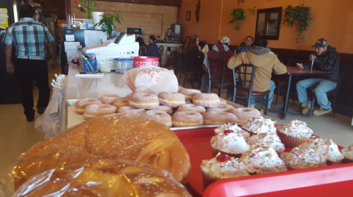 A bakery display with donuts and cupcakes in the foreground, and customers enjoying food in a cozy café setting.