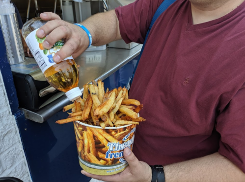 A person in a maroon shirt pours vinegar over a large bucket of French fries.