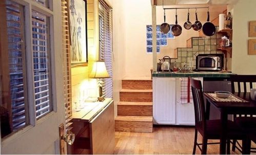 Cozy kitchen with wooden floors, a dining table, and pots hanging above a green-tiled counter. Warm lighting enhances the space.