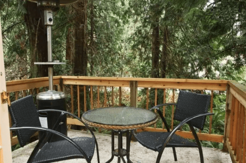 A cozy outdoor seating area with a glass table and two chairs, surrounded by lush greenery.