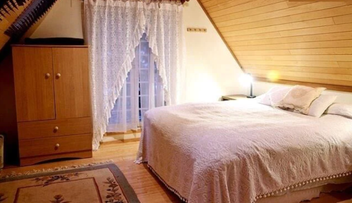 Cozy attic bedroom with a large bed, wooden furniture, and a window draped with lace curtains. Warm, inviting atmosphere.