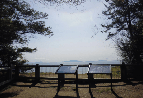 Scenic viewpoint with informational signs overlooking a calm body of water and distant islands, framed by trees.