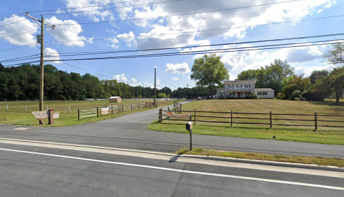 A rural scene featuring a house, grassy fields, and a clear blue sky with scattered clouds. Power lines run overhead.