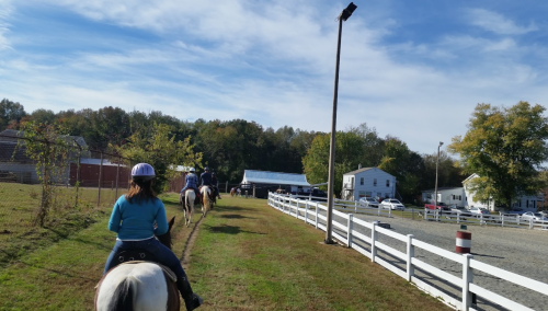 Two people riding horses along a grassy path near a white fence, with buildings and trees in the background.