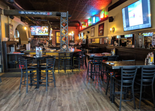 Interior of a lively bar with wooden floors, high tables, colorful lights, and multiple TVs showing sports.