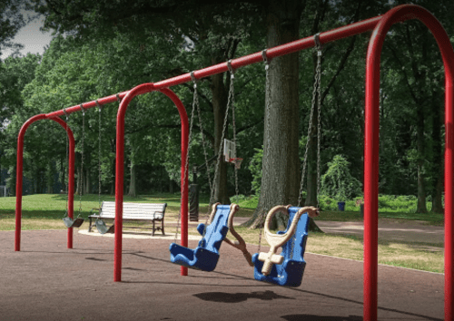 Empty playground swings in a park, surrounded by trees and a bench in the background.