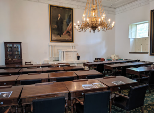 A spacious room with wooden desks, a large chandelier, and a portrait on the wall, featuring a fireplace and windows.