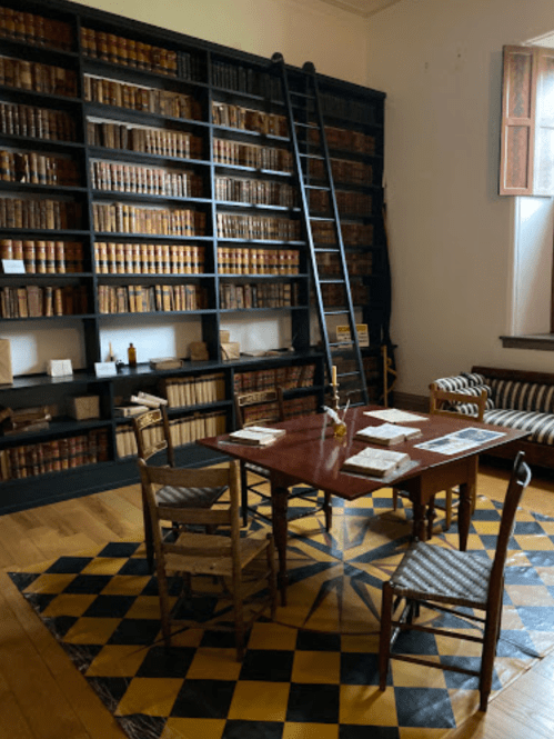A cozy library with a tall bookshelf, a wooden table, chairs, and a patterned rug, featuring warm lighting.