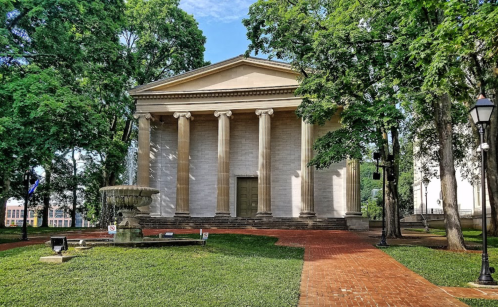 A classical building with columns surrounded by trees and a fountain in a park setting.