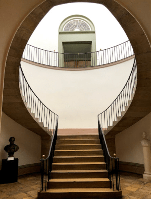 A curved staircase leads to a large window, framed by a smooth, white wall and decorative railings.