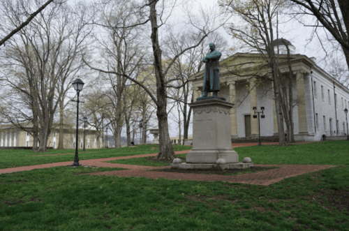 A statue of a figure stands on a pedestal in a park, surrounded by trees and a historic building in the background.