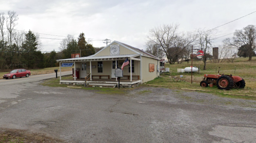 A small country store with a porch, an American flag, a red tractor, and a rural landscape in the background.