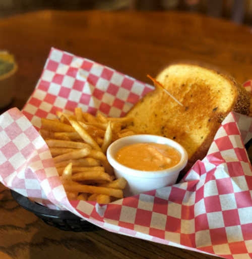 A basket with golden fries, a slice of toasted bread, and a small cup of dipping sauce, all lined with red-checkered paper.