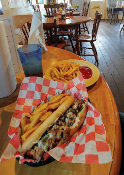 A sandwich with fries in a basket on a wooden table, with a plate of fries and ketchup in the background.