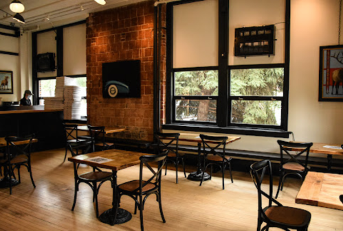 Cozy café interior with wooden tables, black chairs, and large windows overlooking greenery.
