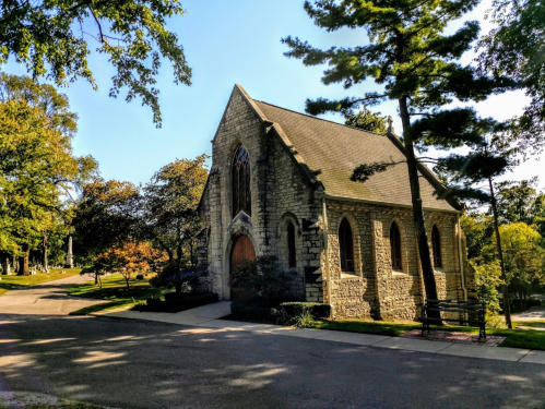 A stone chapel surrounded by trees on a sunny day, with a pathway leading up to it.