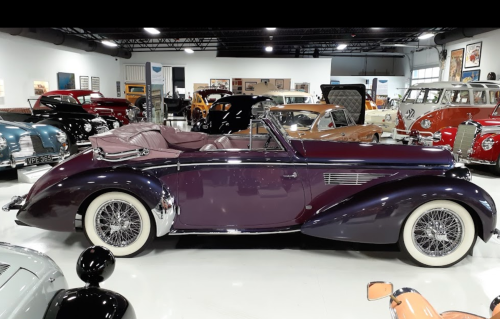 A vintage purple convertible car displayed in a classic car showroom, surrounded by various other antique vehicles.