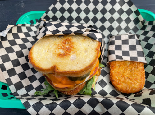 A grilled sandwich with melted cheese and pickles, served with a crispy hash brown, all in checkered paper.