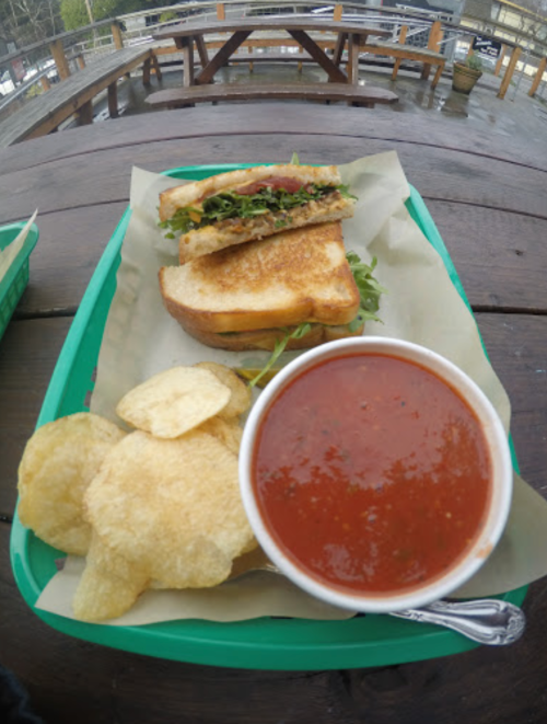 A green tray holds a grilled sandwich with lettuce, a bowl of tomato soup, and a side of potato chips on a wooden table.