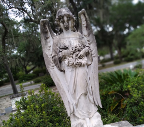 A stone angel statue holding flowers, surrounded by greenery in a serene outdoor setting.
