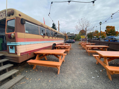A colorful food truck parked beside orange picnic tables in an outdoor dining area, surrounded by trees and cloudy skies.