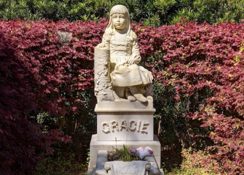 A stone statue of a girl named Gracie sitting on a pedestal, surrounded by vibrant red foliage.