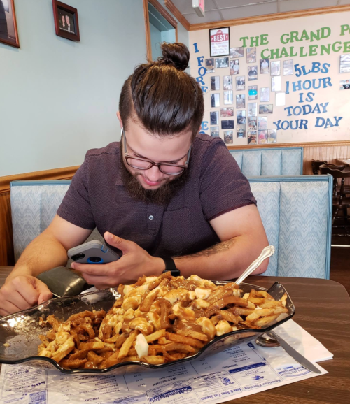 A man with a bun and beard sits at a table, looking at his phone beside a large plate of fries and gravy.